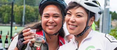 Two women with helmets and jerseys smile and raise small tasting glasses in a toast.